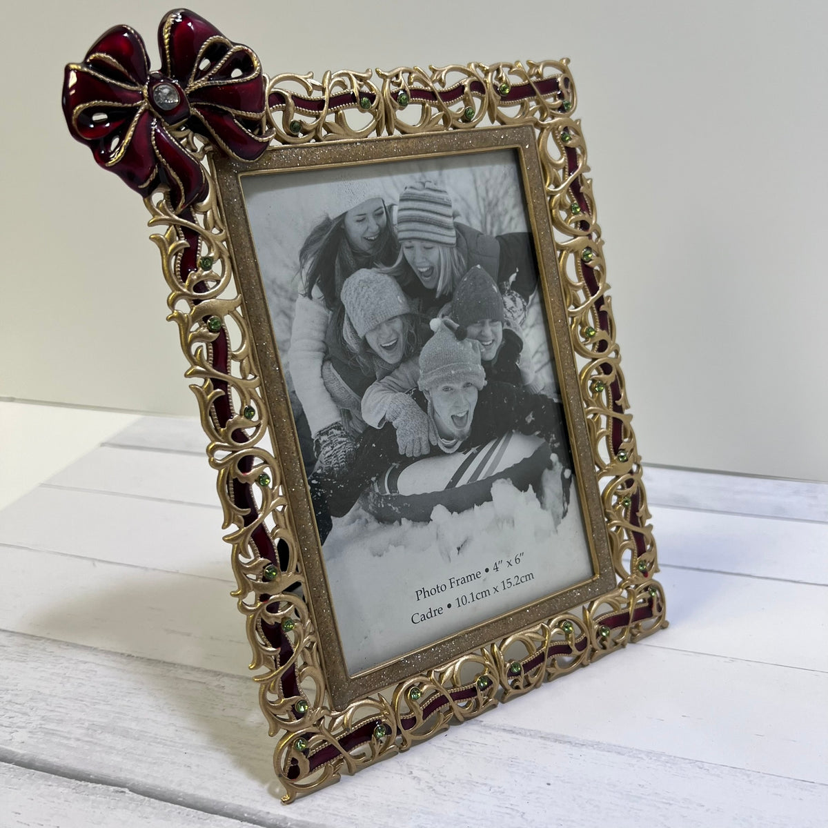 An ornate gold picture frame with a decorative red bow on the top left corner, holding a black-and-white family photo. The frame is shown standing on a white wooden surface against a light gray background.