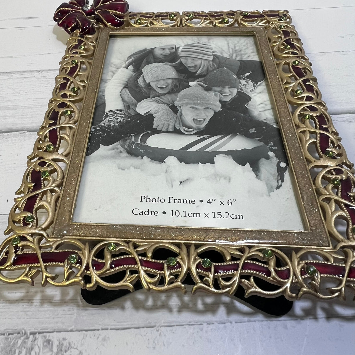 An ornate 4x6 photo frame with an intricate gold filigree border, deep red ribbon accents, and small green crystal embellishments. The frame is shown holding a black-and-white photo of a group of children in the snow, with text printed on the insert indicating the dimensions.
