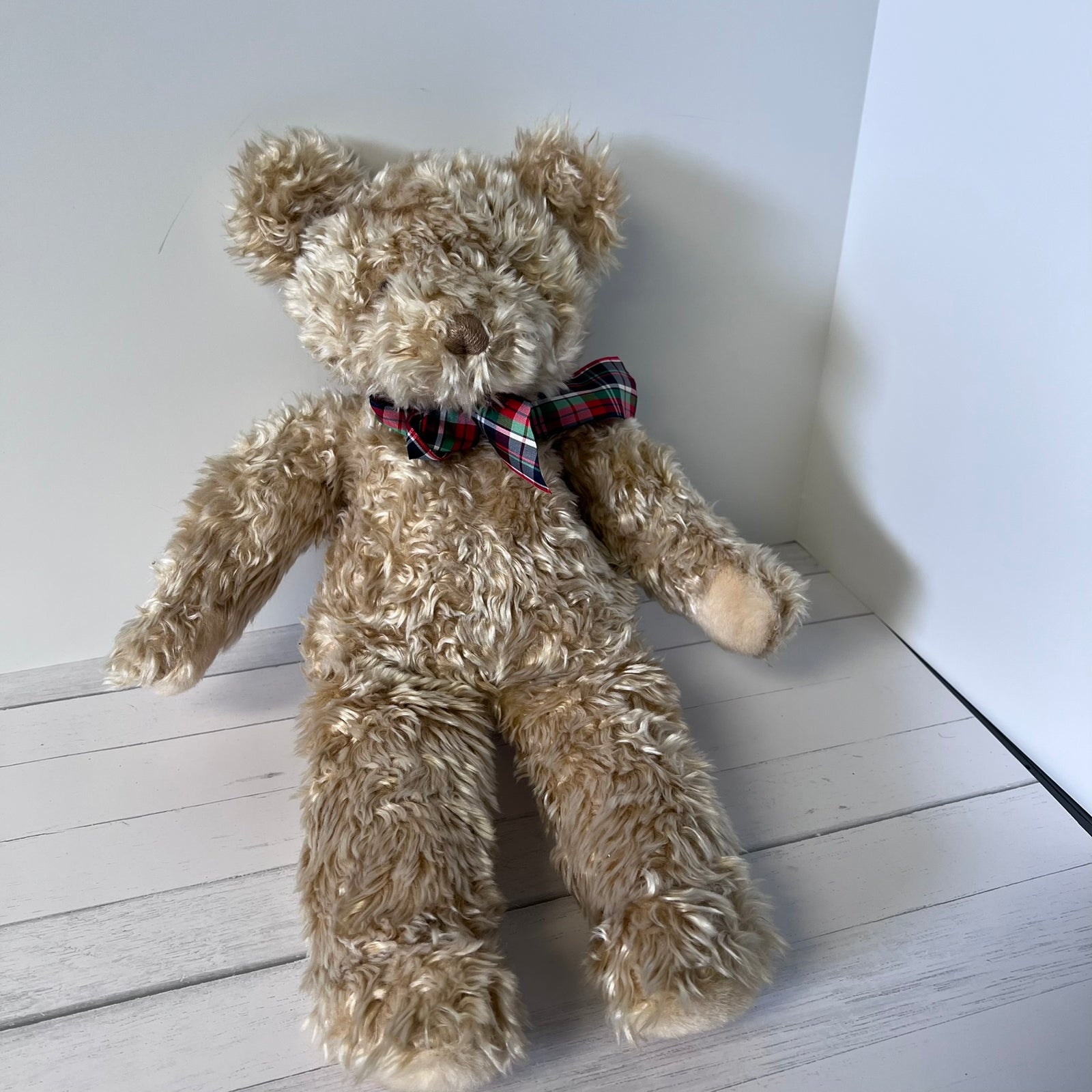 Beige teddy bear with a plaid bow tie on a white wooden floor.