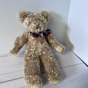 Beige teddy bear with a plaid bow tie on a white wooden floor.