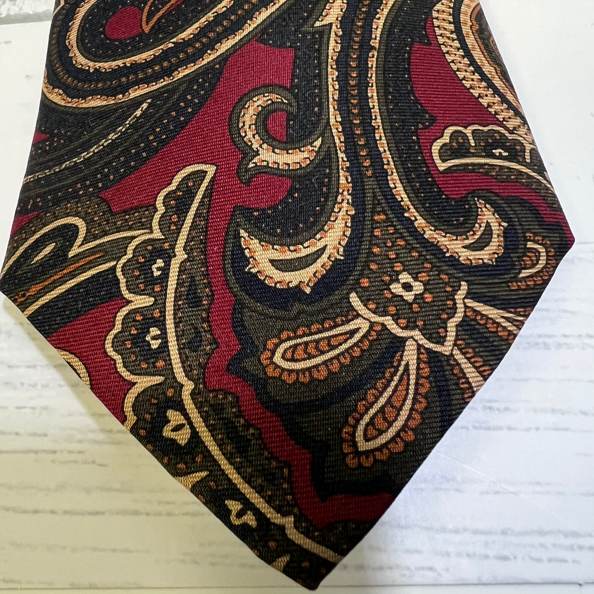 A close-up of a folded necktie featuring a detailed paisley pattern in burgundy red, black, beige, and brown, displayed on a white wood background.