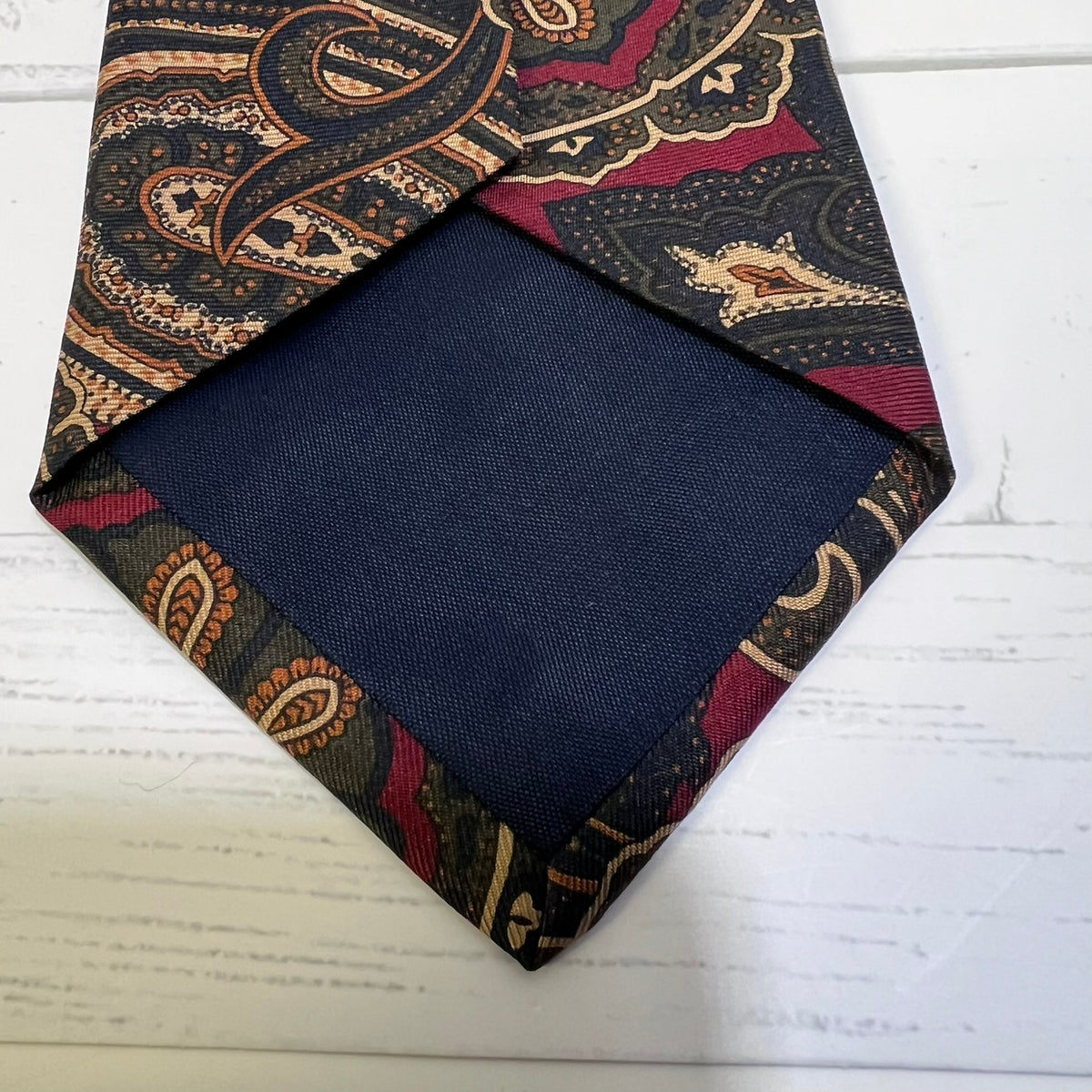 A folded necktie with a navy blue center and a paisley patterned border in black, burgundy, and beige, displayed on a white wood background.