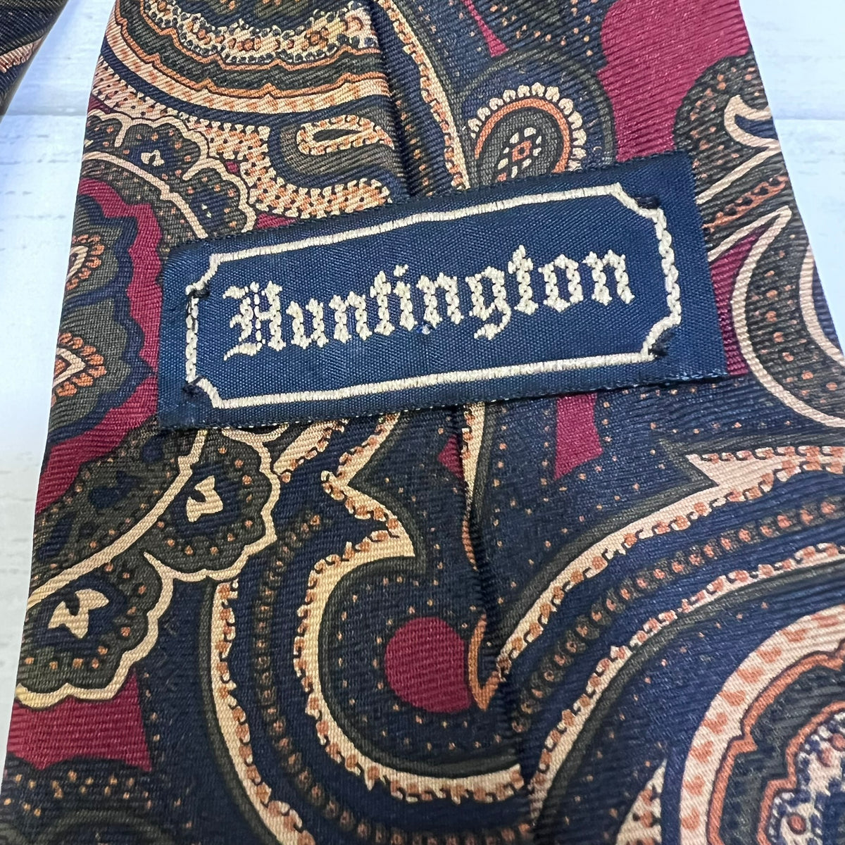 A close-up of a patterned necktie with a navy blue satin label reading 'Huntington' in white gothic lettering. The tie features an intricate paisley pattern in navy, burgundy, beige, and gold.