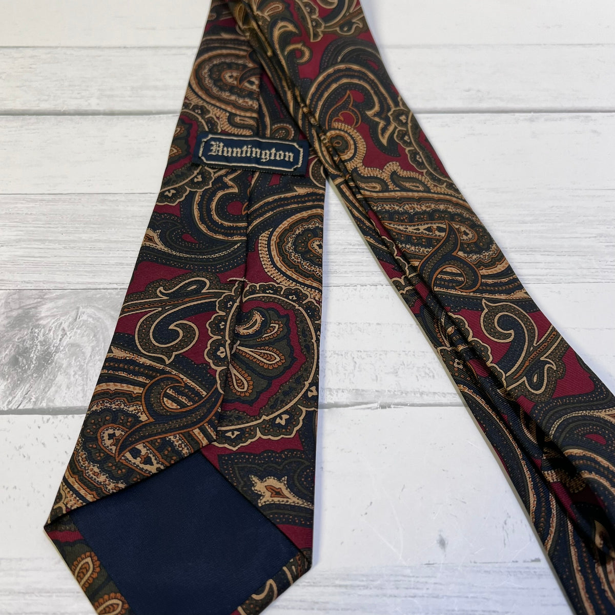 A patterned necktie with a burgundy, gold, and black paisley design, laid flat on a white wood background. The tie features a visible 'Huntington' brand label and a solid navy blue inner end.