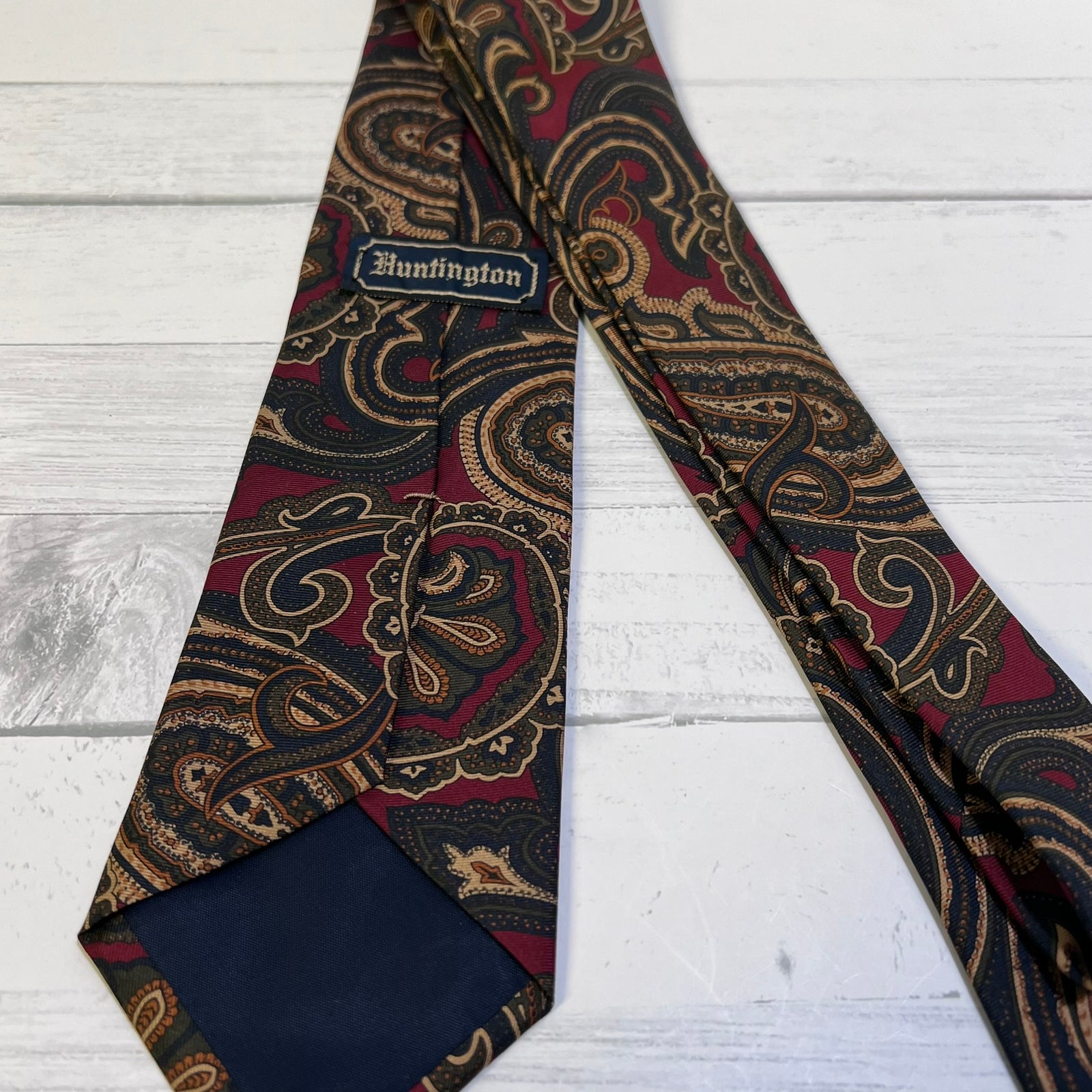 A patterned necktie with a burgundy, gold, and black paisley design, laid flat on a white wood background. The tie features a visible 'Huntington' brand label and a solid navy blue inner end.