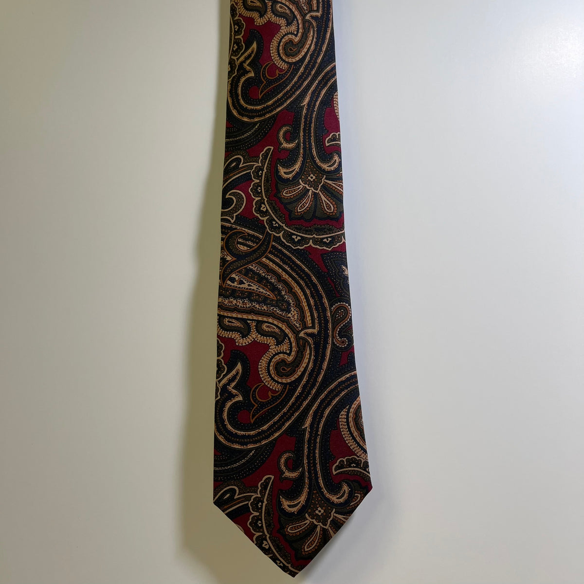 A necktie with an intricate paisley pattern in black, deep red, and gold, hanging against a plain off-white wall.