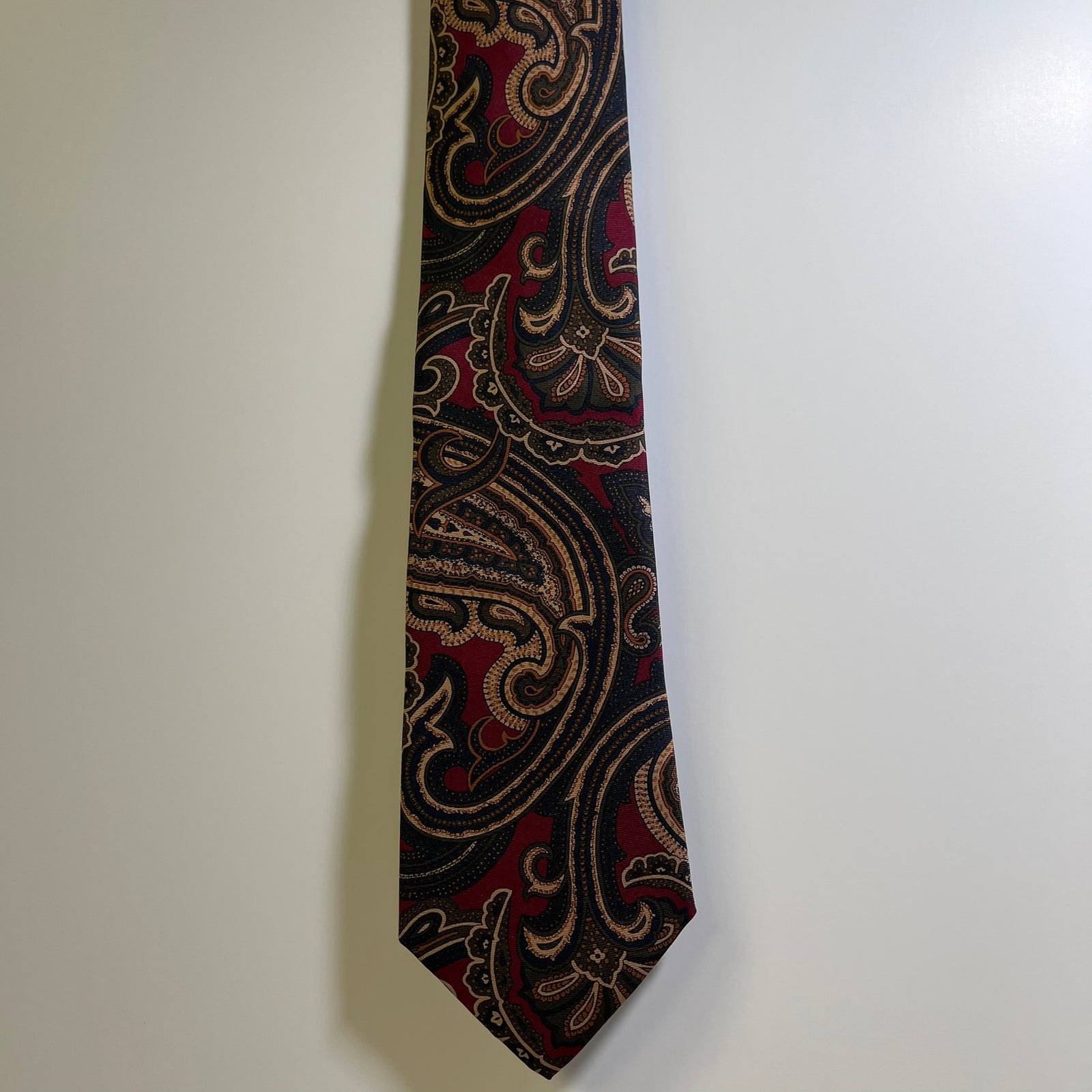 A necktie with an intricate paisley pattern in black, deep red, and gold, hanging against a plain off-white wall.