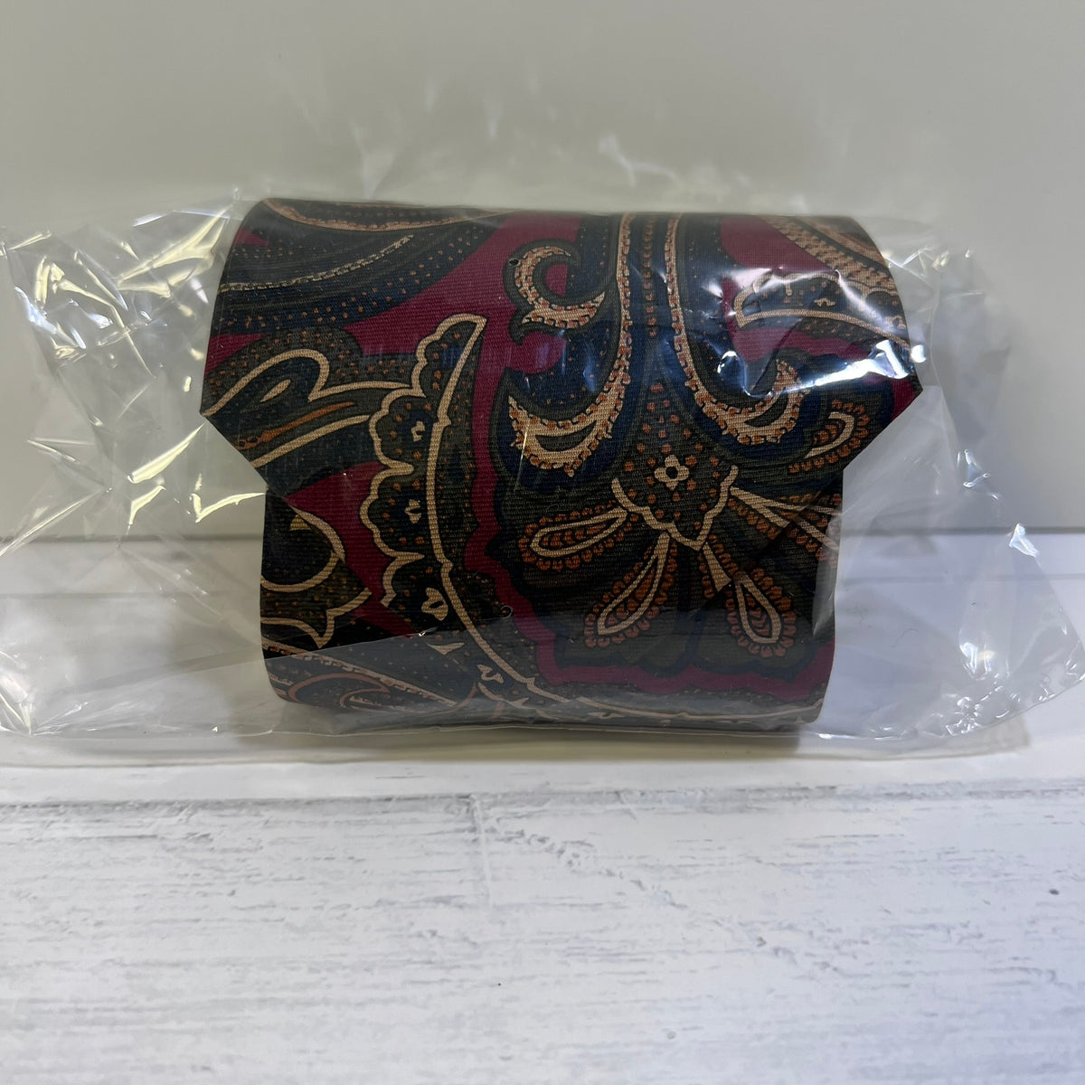 A roll of patterned toilet paper wrapped in clear plastic packaging, featuring a dark paisley design with burgundy, black, and gold details, set against a light gray background.