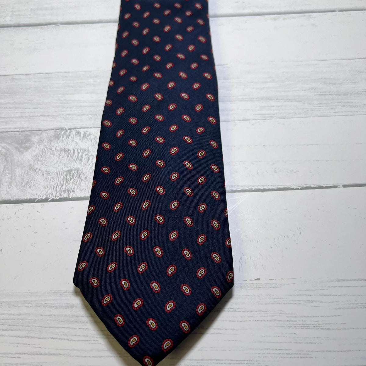 A navy blue necktie with a repeating red and gold paisley pattern, laid flat on a white wood plank surface.