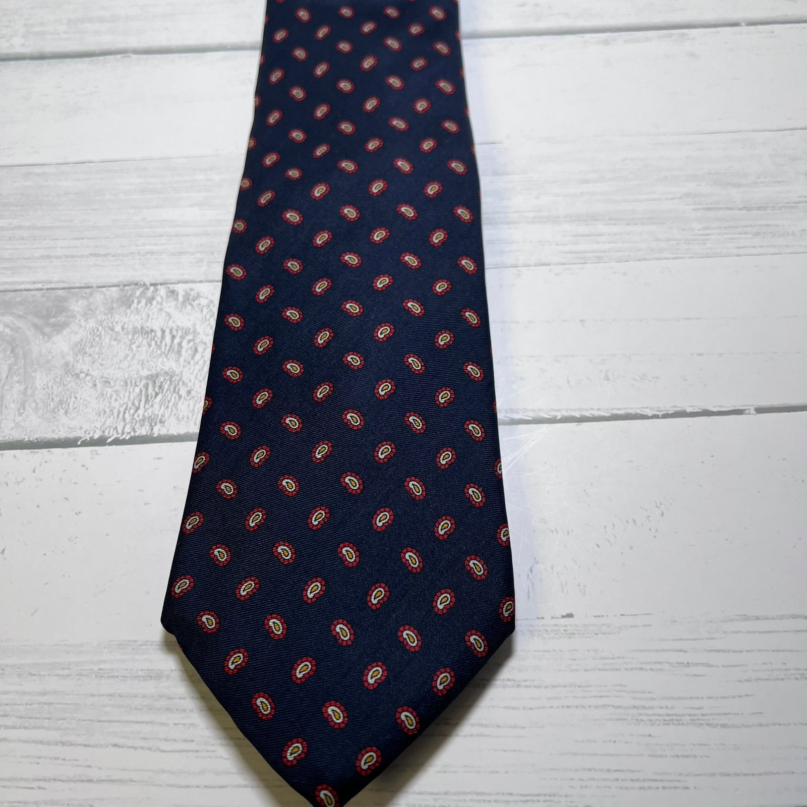 A navy blue necktie with a repeating red and gold paisley pattern, laid flat on a white wood plank surface.