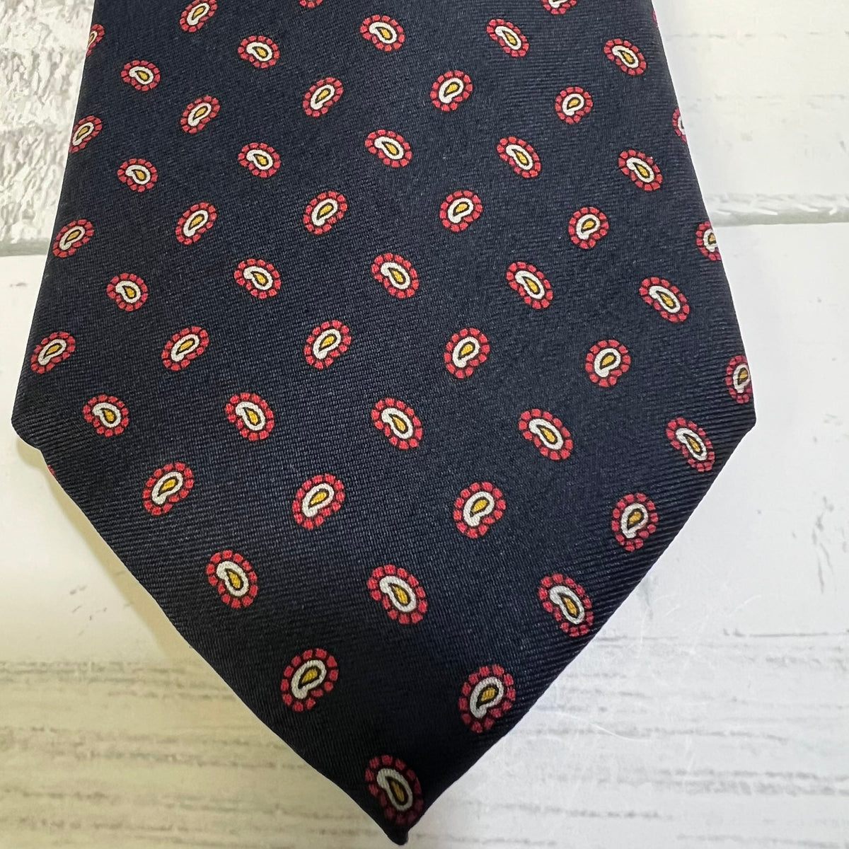 A dark navy necktie with a repeating red, yellow, and white paisley pattern, laid flat on a white wood background.