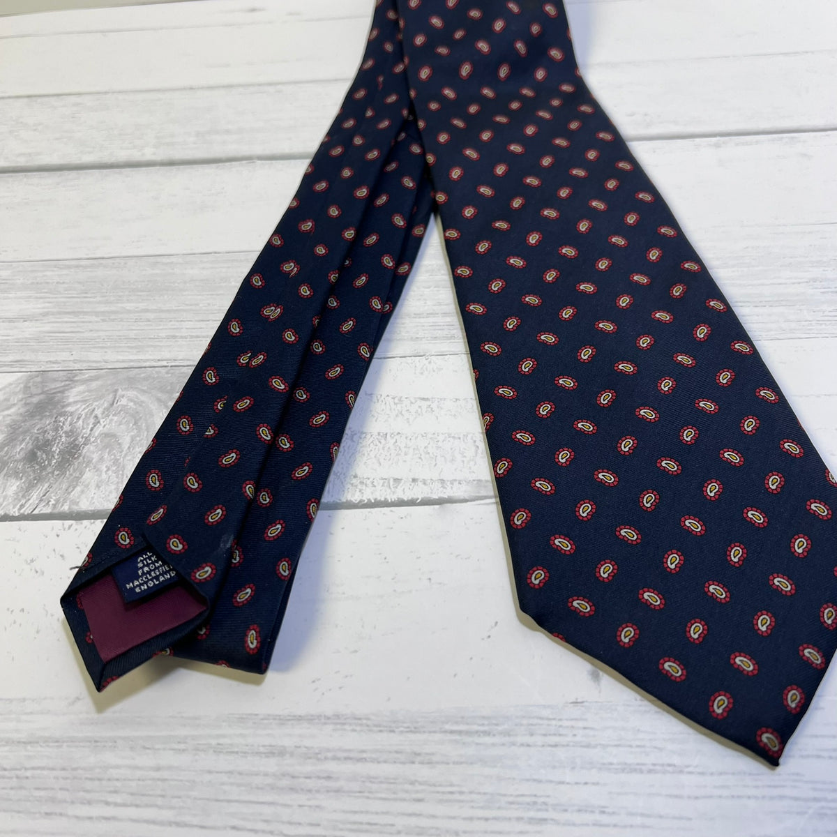 A navy blue necktie with a repeating red and gold paisley pattern, laid flat on a white wood plank surface.