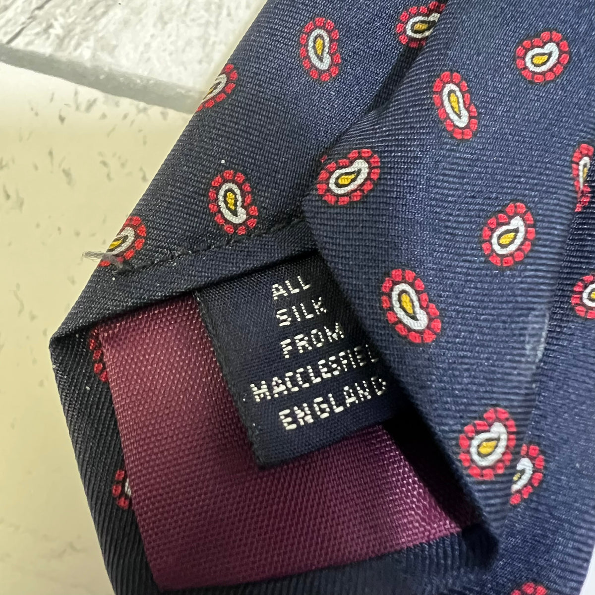 Close-up of a navy blue silk necktie with a red, yellow, and white paisley pattern, featuring a dark label that reads 'ALL SILK FROM MACLESEITE ENGLAND'.