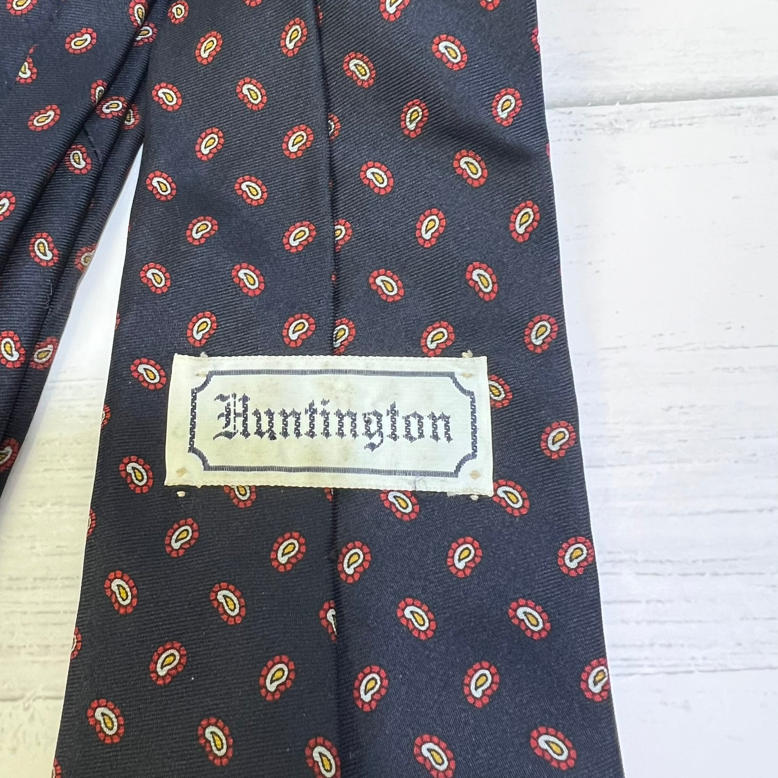 A close-up of a patterned necktie with a visible Hunterston label, featuring a dark navy base with small red, yellow, and white paisley-like motifs, laid flat on a white wood surface.