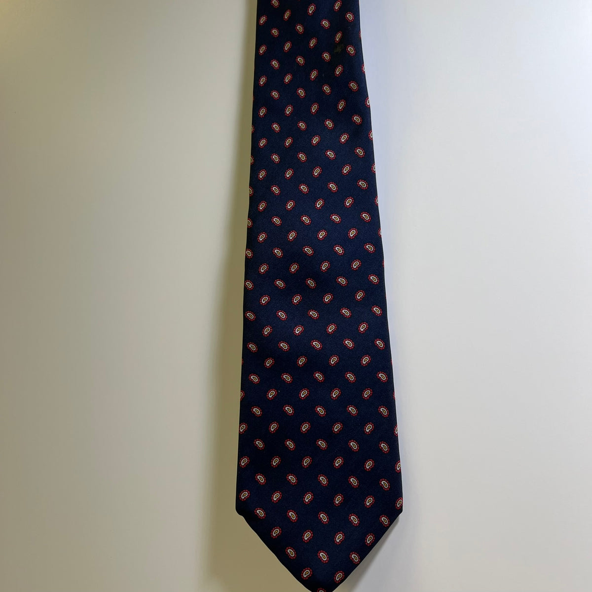 A dark navy blue necktie with a repeating small red and white paisley pattern, hanging against a plain off-white wall.