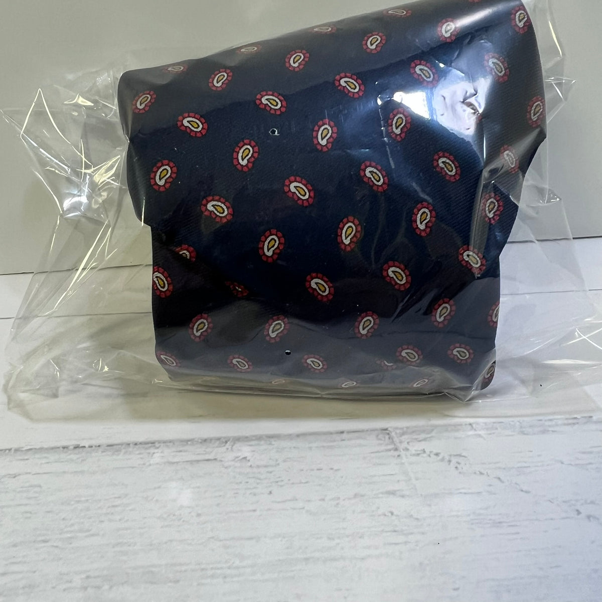 A dark navy blue fabric item, likely a pocket square or small fabric accessory, featuring a repeating red, white, and beige paisley pattern, wrapped in clear plastic packaging on a white wooden surface.