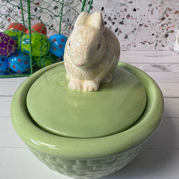 Green ceramic bowl with a rabbit-shaped lid on a wooden surface, colorful eggs in the background.