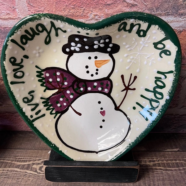 Heart-shaped decorative plate with a snowman design and text on a wooden surface.