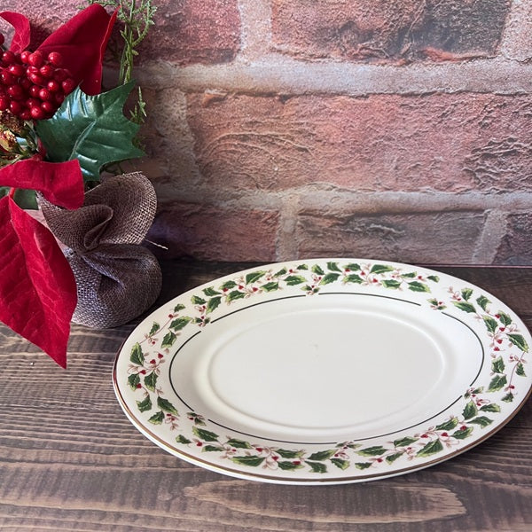 Decorative plate with holly design on a wooden surface next to poinsettias and a brick wall.