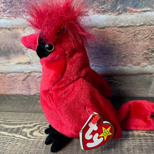 Red plush cardinal toy with a visible tag on a wooden surface and brick wall background