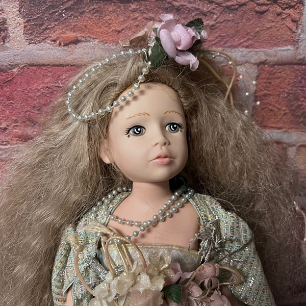 Doll with long hair, floral headband, and pearl necklace against a brick wall.