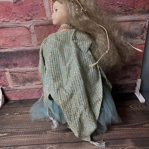 Doll with a green textured garment against a brick wall.