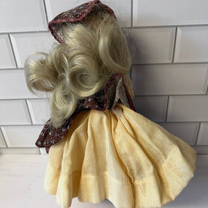 Vintage-style doll with blonde hair and a yellow dress against a tiled wall.