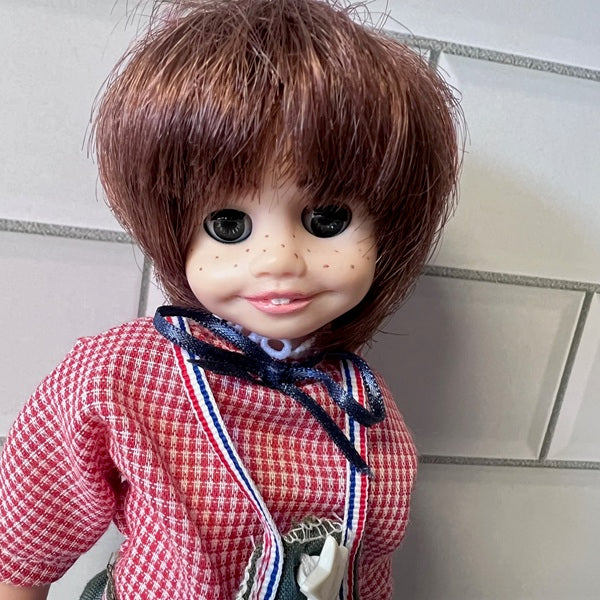  Doll with brown hair and a red checkered shirt against a tiled wall background