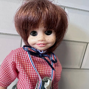  Doll with brown hair and a red checkered shirt against a tiled wall background