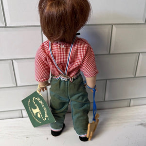 Doll wearing a red checkered shirt, green suspenders, and green pants holding a green book against a tiled wall.