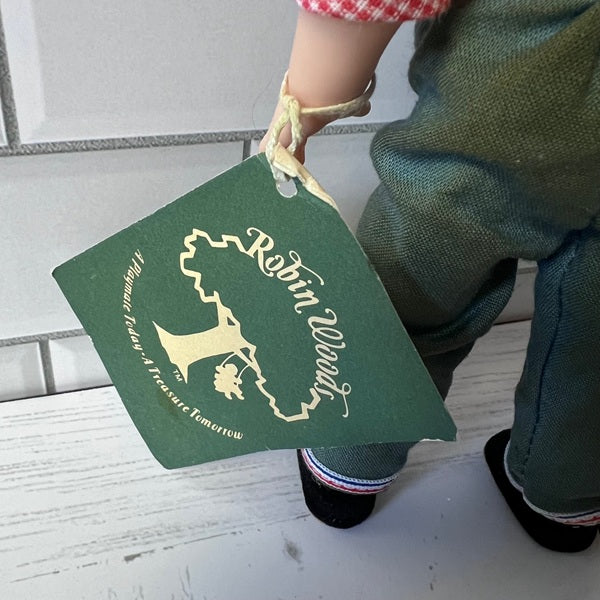 Green tag with gold text and logo held by Robin Woods doll hand on a light wooden floor.