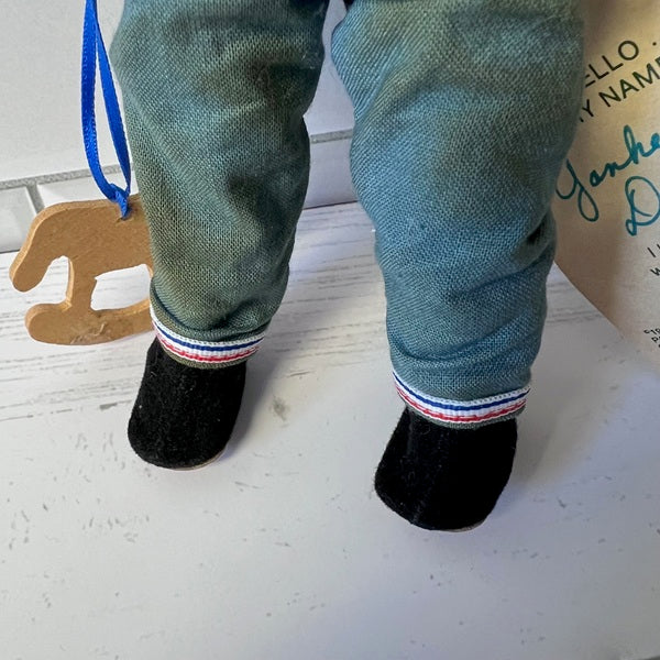 Close-up of a doll wearing green pants with a striped cuff and black socks on a light wooden floor.