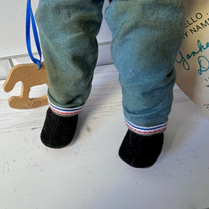 Close-up of a doll wearing green pants with a striped cuff and black socks on a light wooden floor.