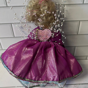 Doll with a purple dress and decorative headpiece against a tiled wall.