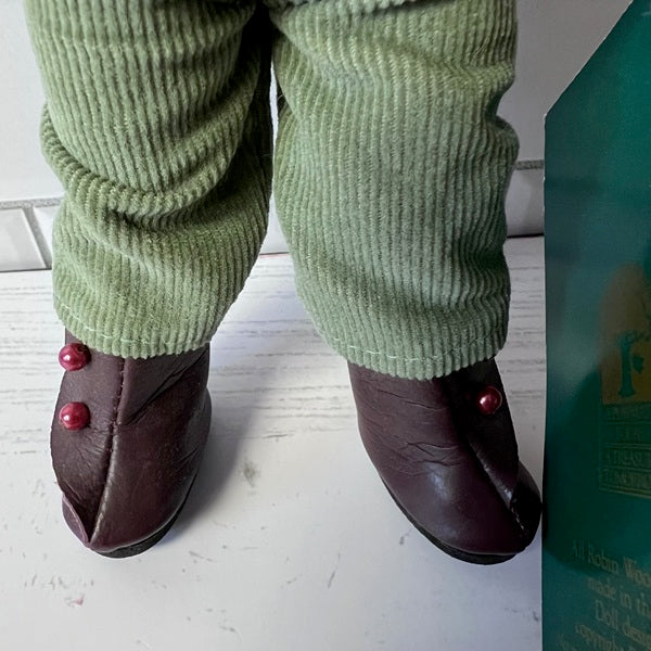Doll wearing green pants and brown shoes with red buttons, standing next to a bottle.