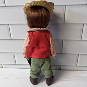 Doll wearing a red jacket, green pants, and straw hat against a white tiled wall.