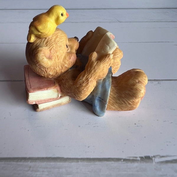 Figurine of a bear reading a book with a chick on its head against a light wooden background