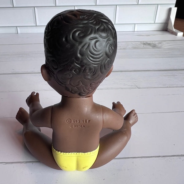 Toy figure of a child with textured hair, wearing a yellow diaper, on a light wooden floor.