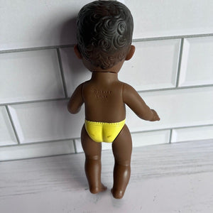 Toy figure of a child in a yellow diaper against a tiled wall.