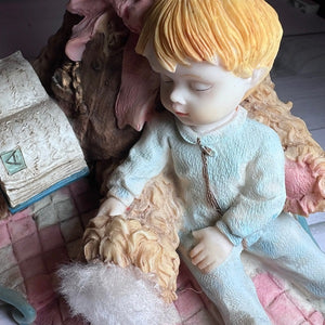 Vintage-style figurine of a child holding a bear, with a book and decorative elements in the background.