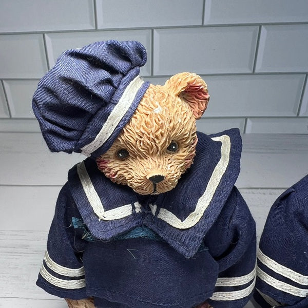 Teddy bear dressed in a navy blue sailor outfit with a white stripe against a light gray tiled wall.