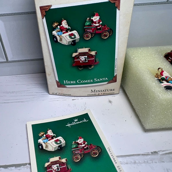 Set of miniature Santa Claus figurines in a 'Here Comes Santa' display box on a white surface.
