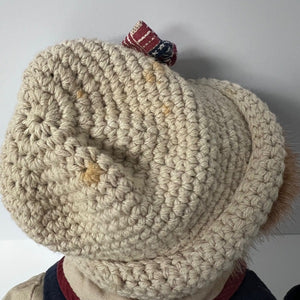 Close-up of a beige knitted beanie with a small decorative item on top against a plain background.