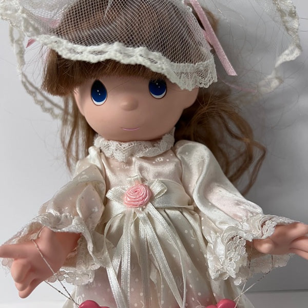 Doll in a white dress with lace details and a pink rose, wearing a matching hat.