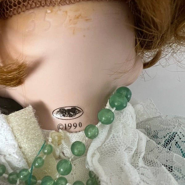 Vintage doll with green beads and white lace, featuring a brand logo and year.