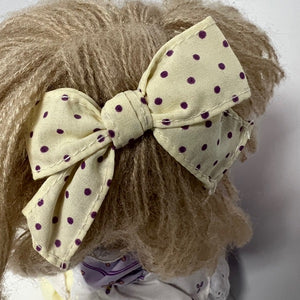 Close-up of a doll with a beige bow featuring purple polka dots on a plain background