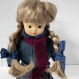 Doll with blonde hair styled in pigtails, wearing a blue outfit and red scarf on a white background