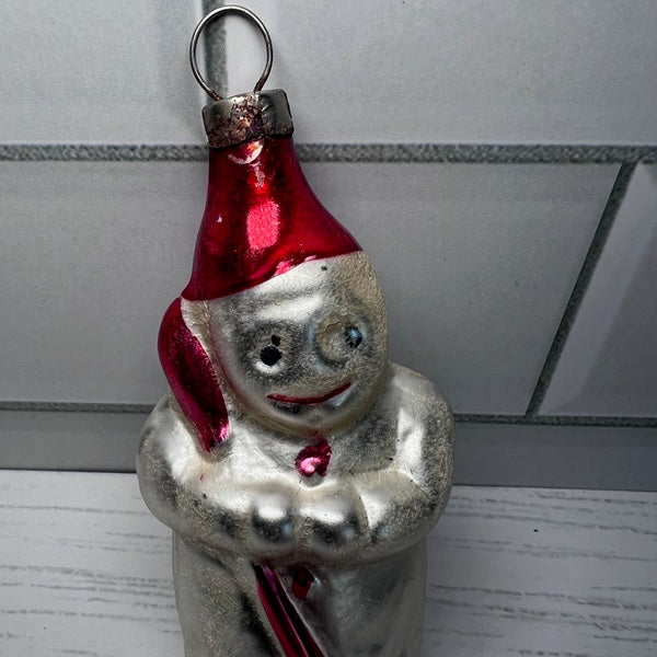 Vintage glass snowman ornament with a red hat and candy cane against a tiled wall background.