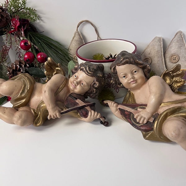 Two cherub figurines playing music with a decorative wreath in the background.
