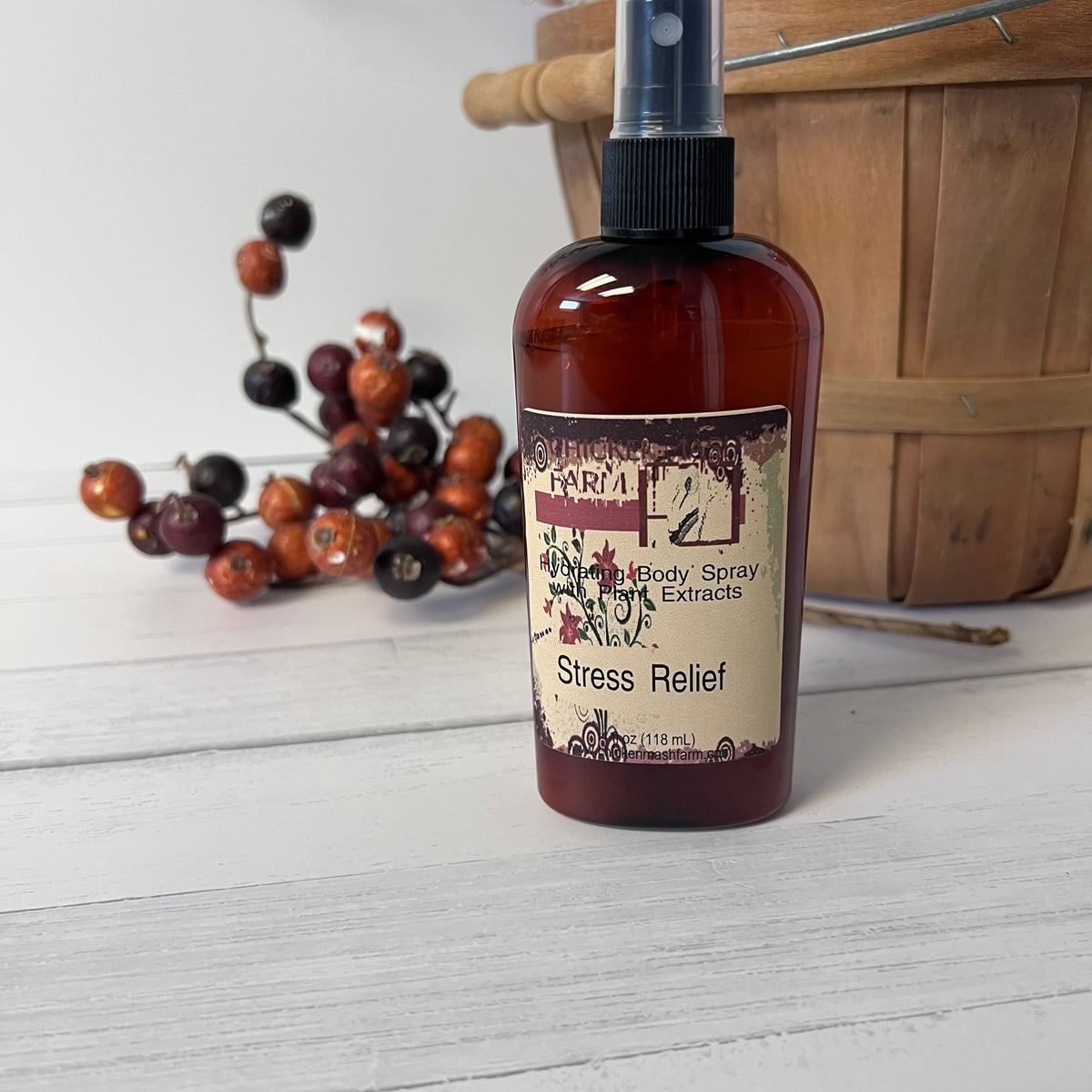 An amber body spray bottle labeled Chickenmash Farm Body Spray Stress Relief  with a black spray top, set on a white wood surface with decorative berries and a wooden basket in the background.